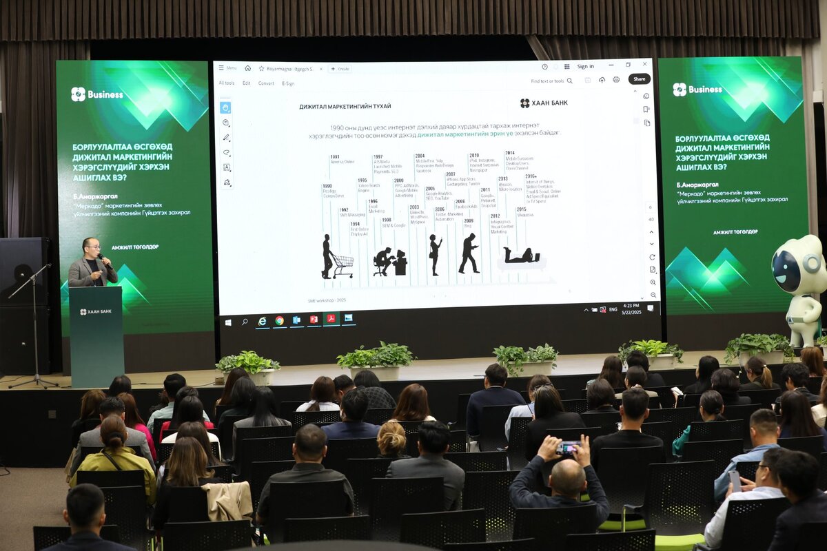 A speaker presents to an audience in a conference hall, showing a large screen with a diagram. Green banners display SME workshop: Бүтээгдэхүүн бус шийдэл санал болго, and a robot stands on the right side of the stage.