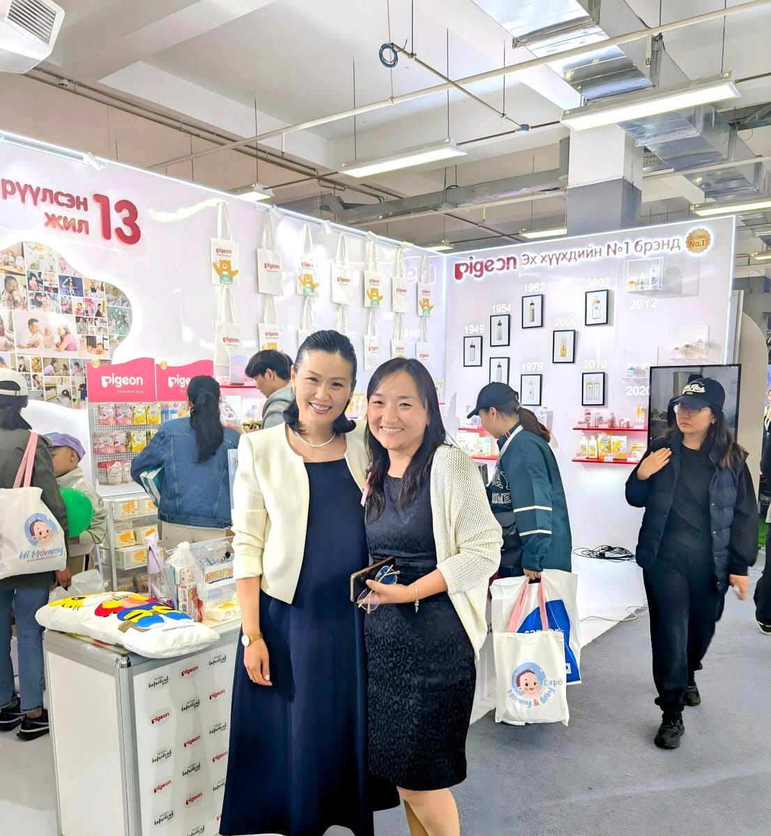 Two women smile and pose together in front of a Pigeon brand booth at a brightly lit indoor event. Other people browse products on display shelves in the background.