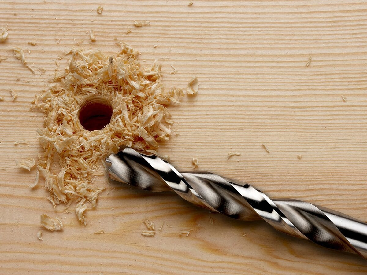 A close-up of a wooden surface with a fresh hole drilled into it, surrounded by wood shavings. A large, silver drill bit rests next to the hole.