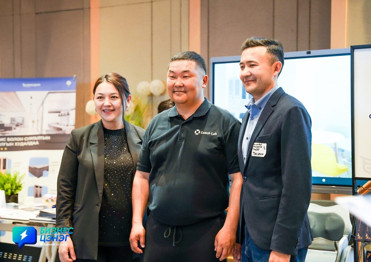 Three people stand together, smiling for a photo at an indoor event. Two men and one woman are wearing business casual clothing. Banners and screens are visible in the background.