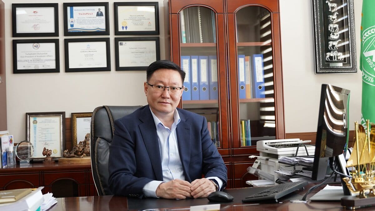 A man in a suit sits at a desk in an office with framed certificates, awards, and shelves of binders behind him. A computer, printer, and office supplies are on the desk.