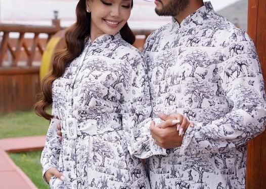 A couple wearing matching white outfits with detailed black animal print patterns and white hats stands holding hands outdoors, smiling at each other. The background features wooden structures and a domed building.