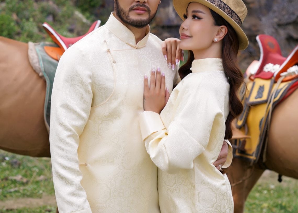 A man and woman in traditional cream-colored attire and straw hats pose together outdoors with horses and rocky terrain in the background. The woman leans on the man, both looking towards the camera.