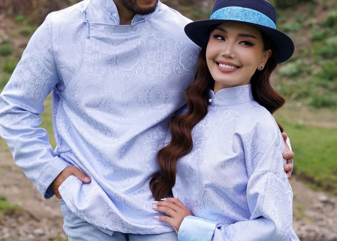 A man and a woman wearing matching light blue traditional shirts and blue hats pose together outdoors with a rocky background. They both have long sleeves and are smiling gently. The image features the Tumen Torgo logo.