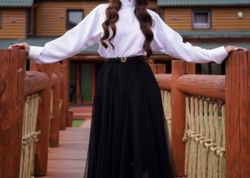 A woman stands on a wooden bridge, wearing a white blouse, black tulle skirt, black hat, and white heels. Behind her is a large wooden building. The image is branded with the Tumen Torgo logo.