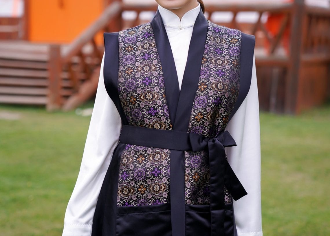 A smiling woman stands outdoors on grass, wearing a traditional Mongolian vest with intricate patterns over a white blouse and black skirt. A round yurt and wooden steps are visible in the background.