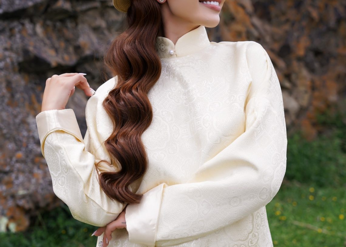 A woman wearing a beige traditional embroidered top and hat stands outdoors in front of a rocky background, smiling and looking to the side, with long wavy hair over her shoulder.