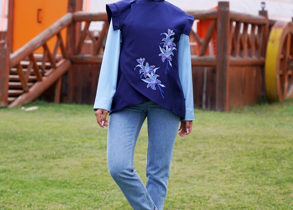 A woman stands on grass in front of a wooden fence and a yurt, wearing blue jeans, white sneakers, and a blue top with decorative floral embroidery. The Tumen Torgo logo appears in two corners.