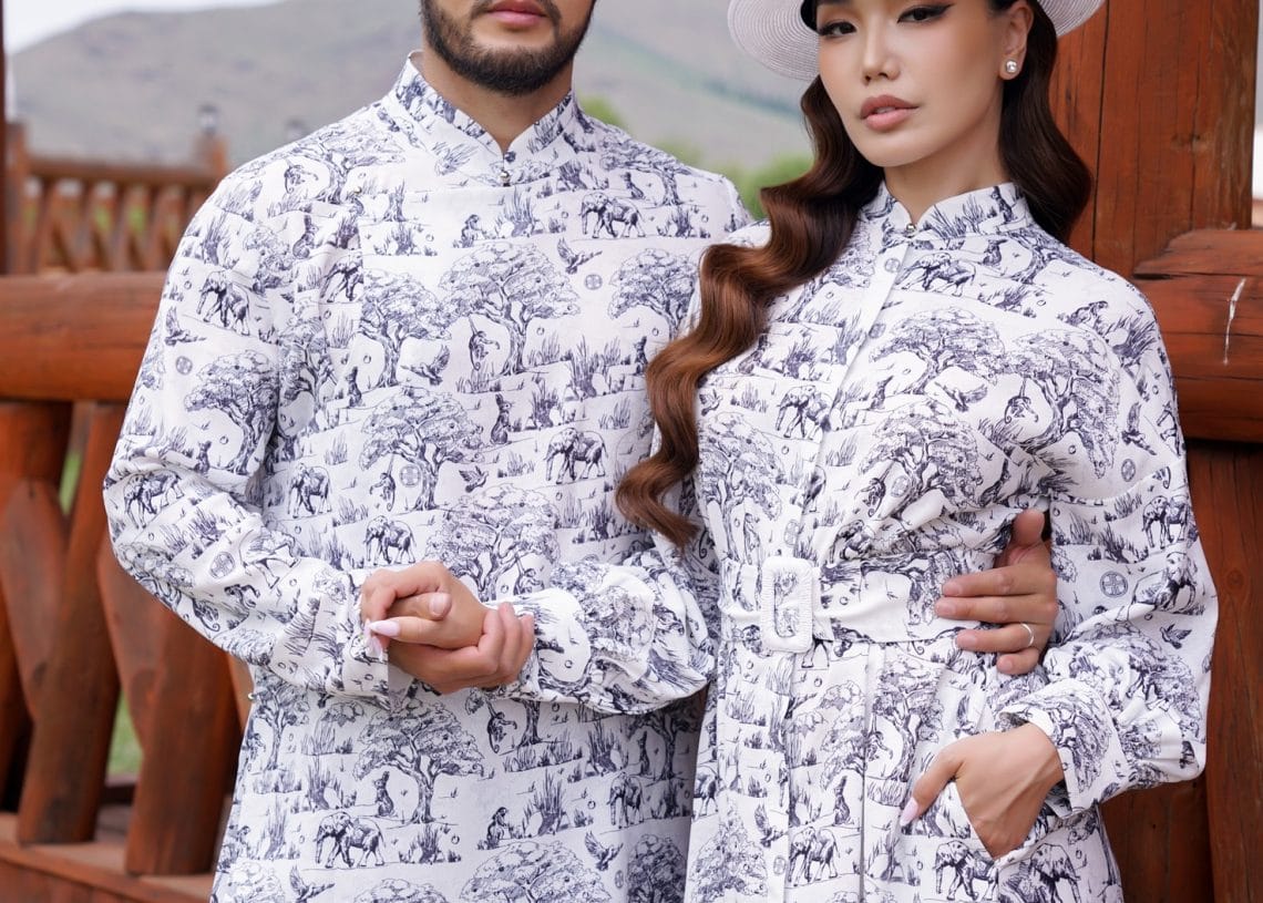 A man and woman stand close together outdoors, both wearing matching white outfits with black animal prints and white hats. Wooden fencing and hills are visible in the background. Tumen Torgo logo is in the corners.