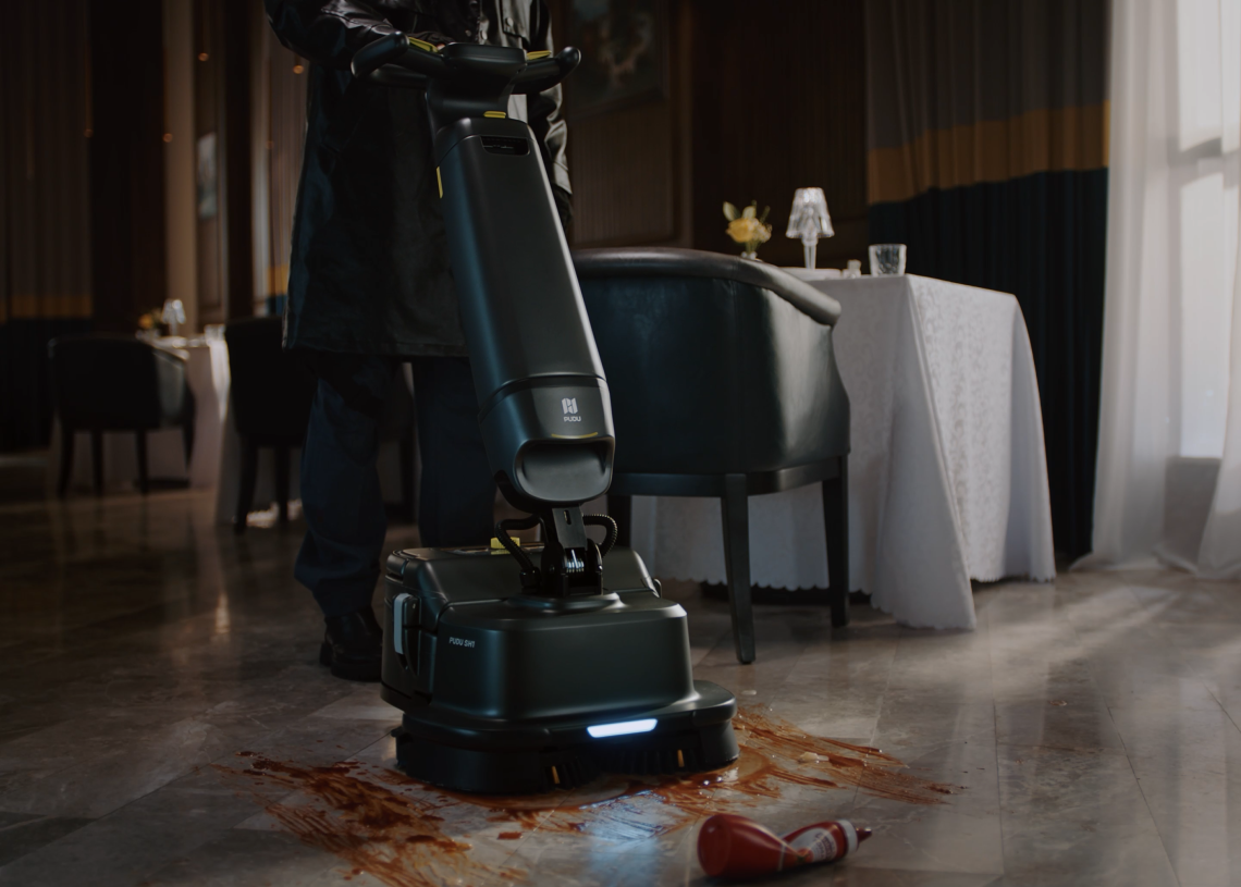 A person using a floor cleaning machine to clean a red liquid spill and a fallen cup on a marble floor in a dimly lit restaurant setting.