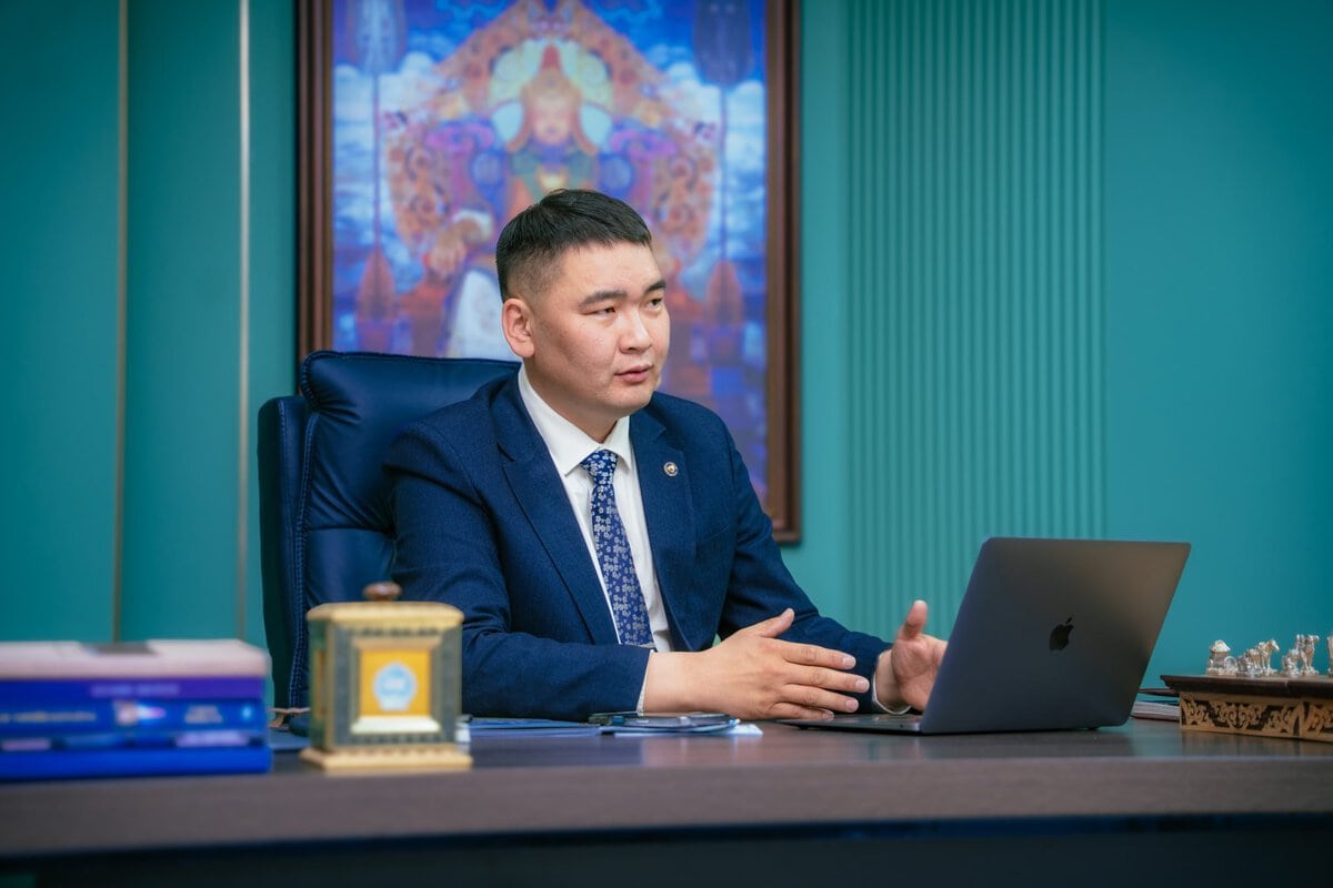 A man in a blue suit sits at a desk with a laptop, gesturing as he speaks. Behind him is a colorful painting with intricate patterns, and books and decorative items are on the desk.