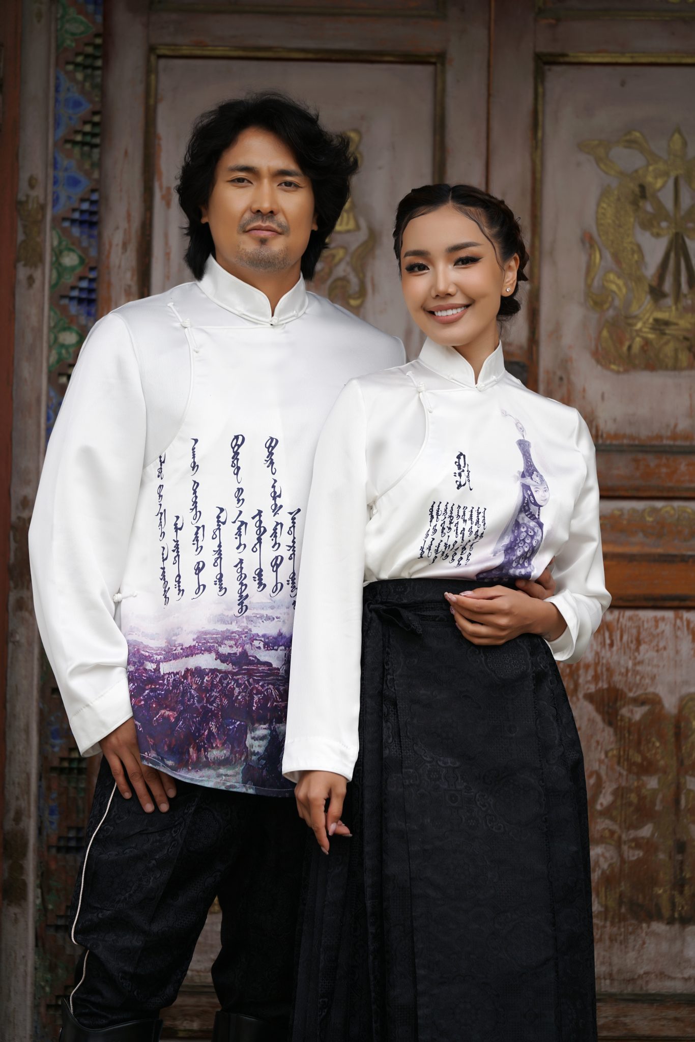 A man and woman stand side by side wearing traditional Mongolian attire with white tops featuring script and artistic designs, posing in front of a wooden door with ornate details and carvings.