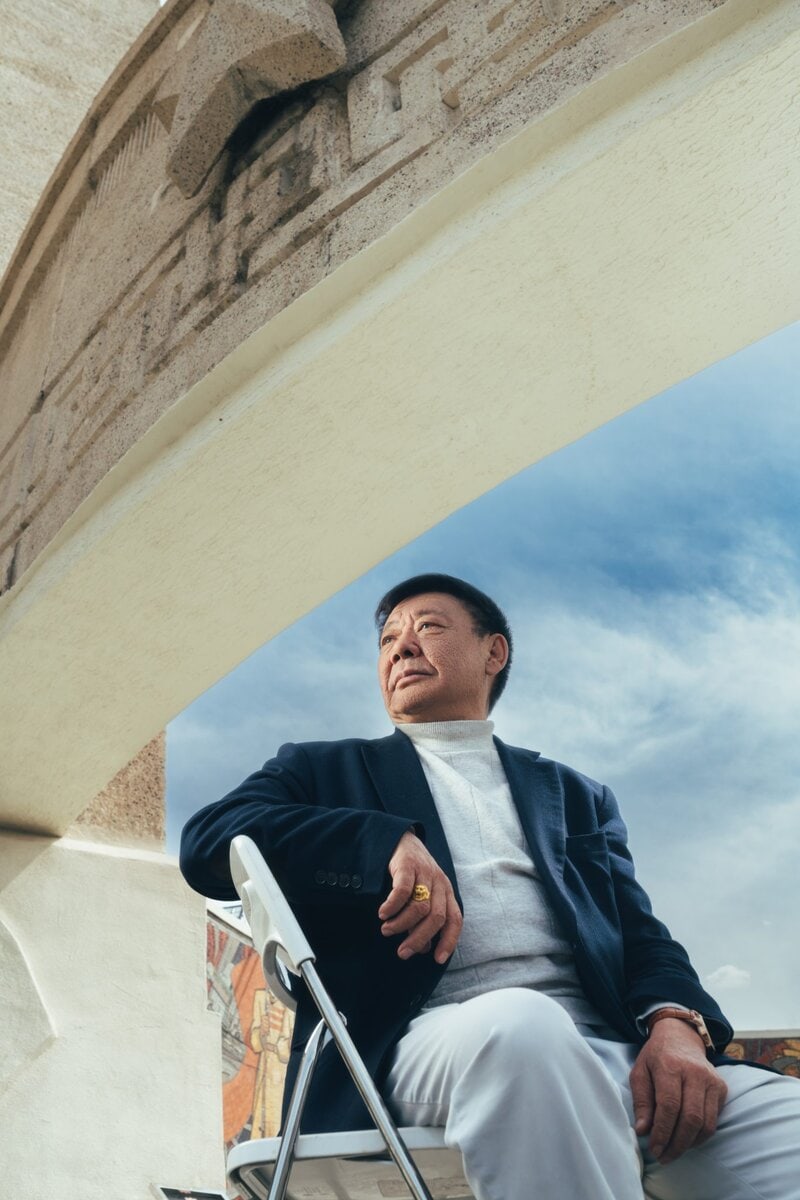 A man in a dark blazer and white pants sits on a chair outside under a stone arch, looking into the distance with a thoughtful expression. The sky is partly cloudy in the background.
