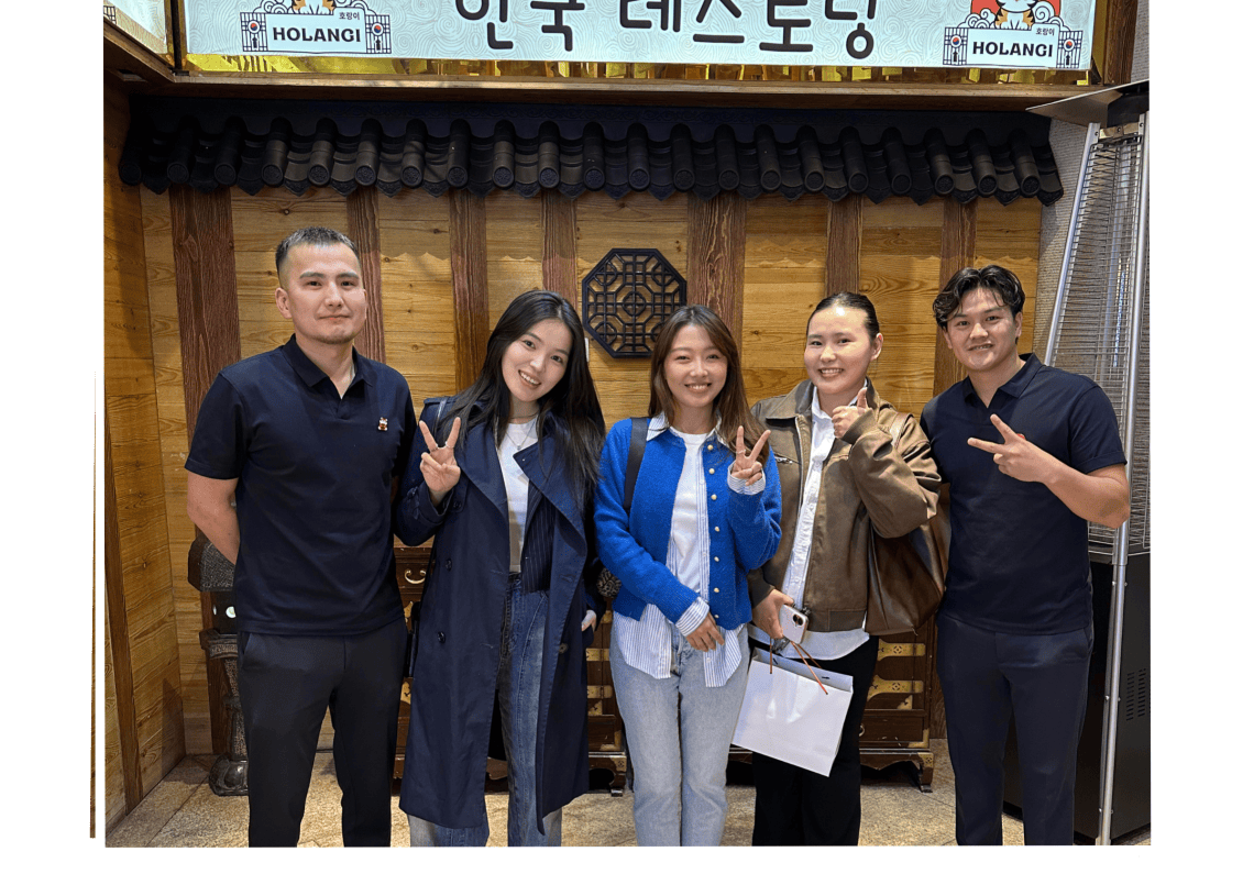 Five people stand smiling and posing with peace signs in front of a Korean restaurant entrance with a wooden facade and a sign written in Korean above them.