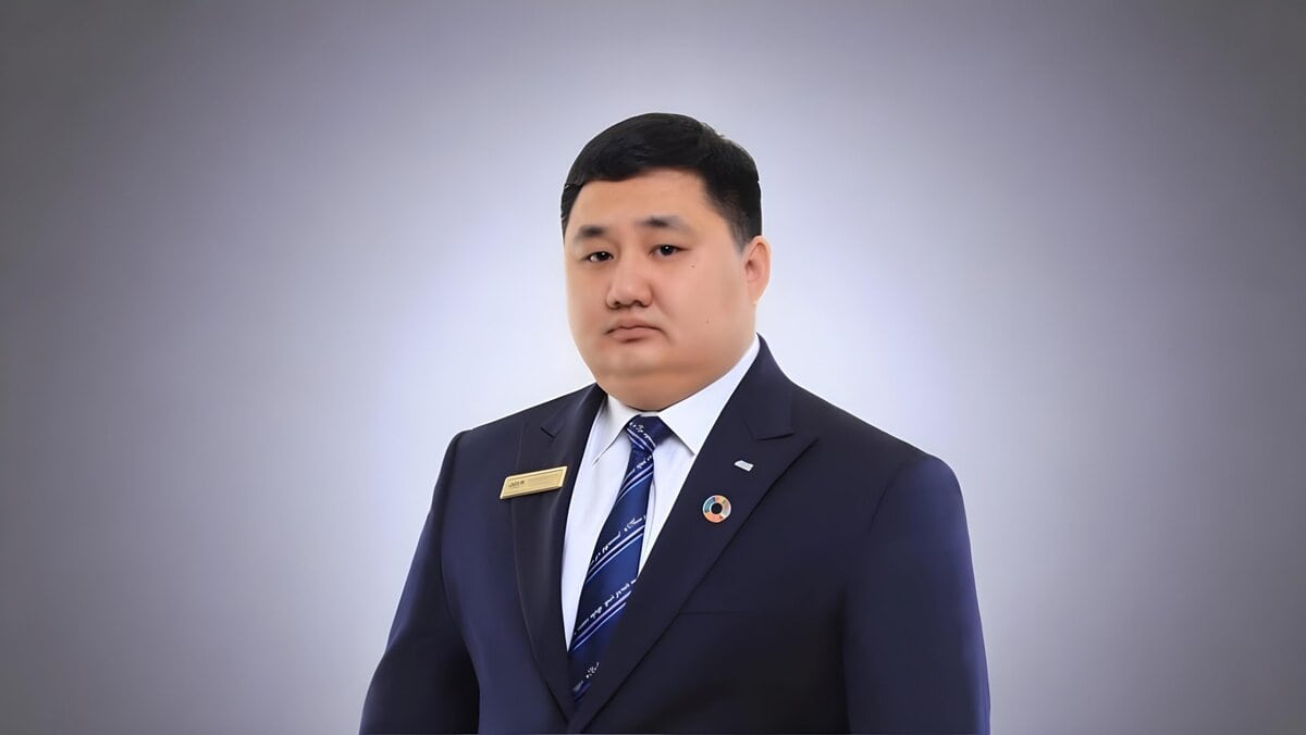 A man in a dark suit and striped tie stands against a plain, gradient background. He wears a name badge and a lapel pin, looking directly at the camera with a neutral expression.