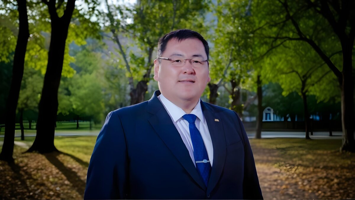 A man wearing glasses and a dark blue suit with a blue tie stands outdoors in a park with green trees and sunlight filtering through the leaves.