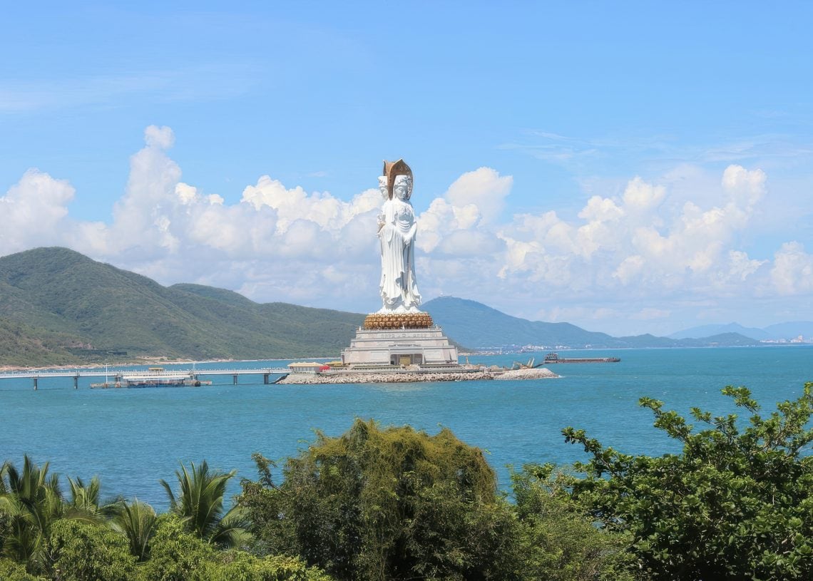 A large white statue of the Guanyin of Nanshan stands on an island in the sea, connected by a bridge, with green hills, blue sky, and scattered clouds in the background. Trees frame the foreground.