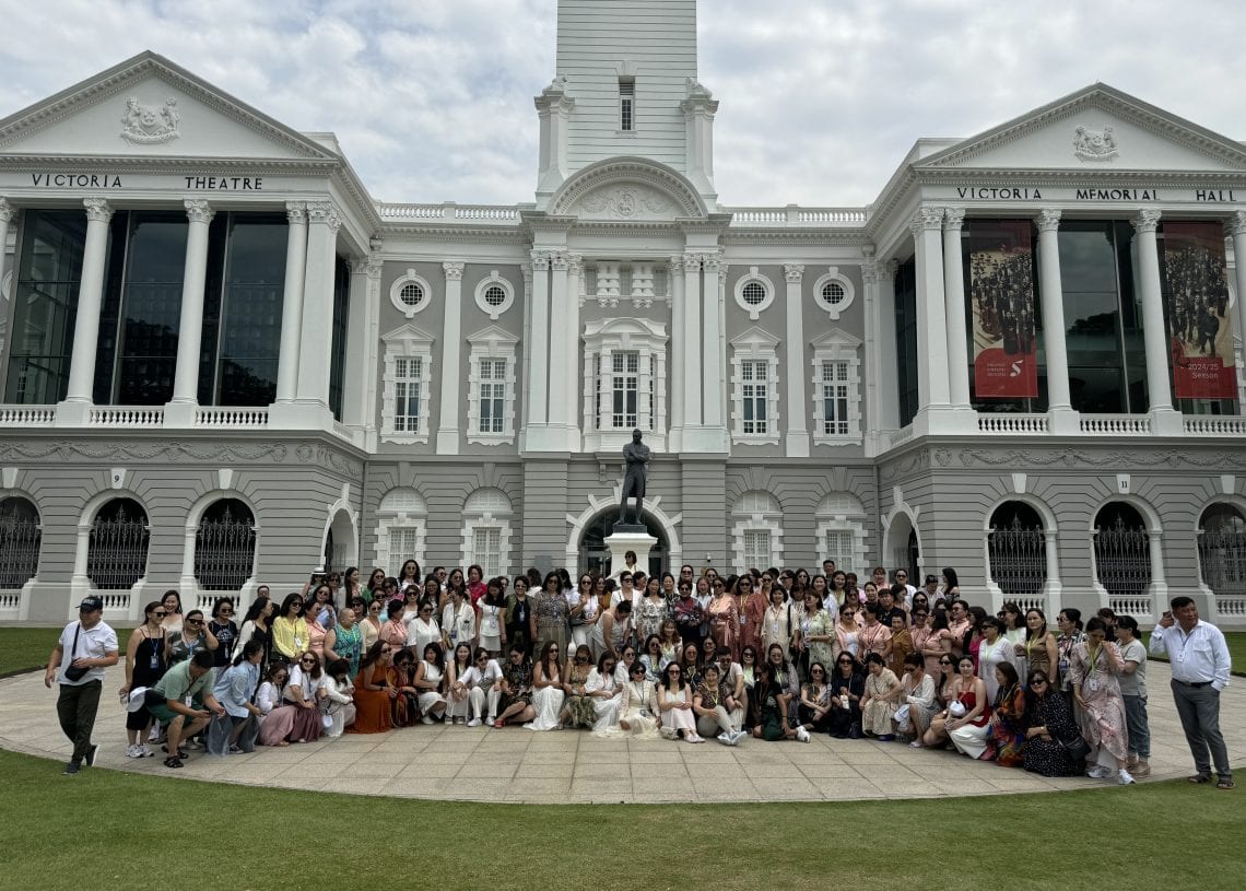 A large group of people pose for a photo in front of the Victoria Theatre and Concert Hall, a historic white building with columns and arched windows, under a cloudy sky.