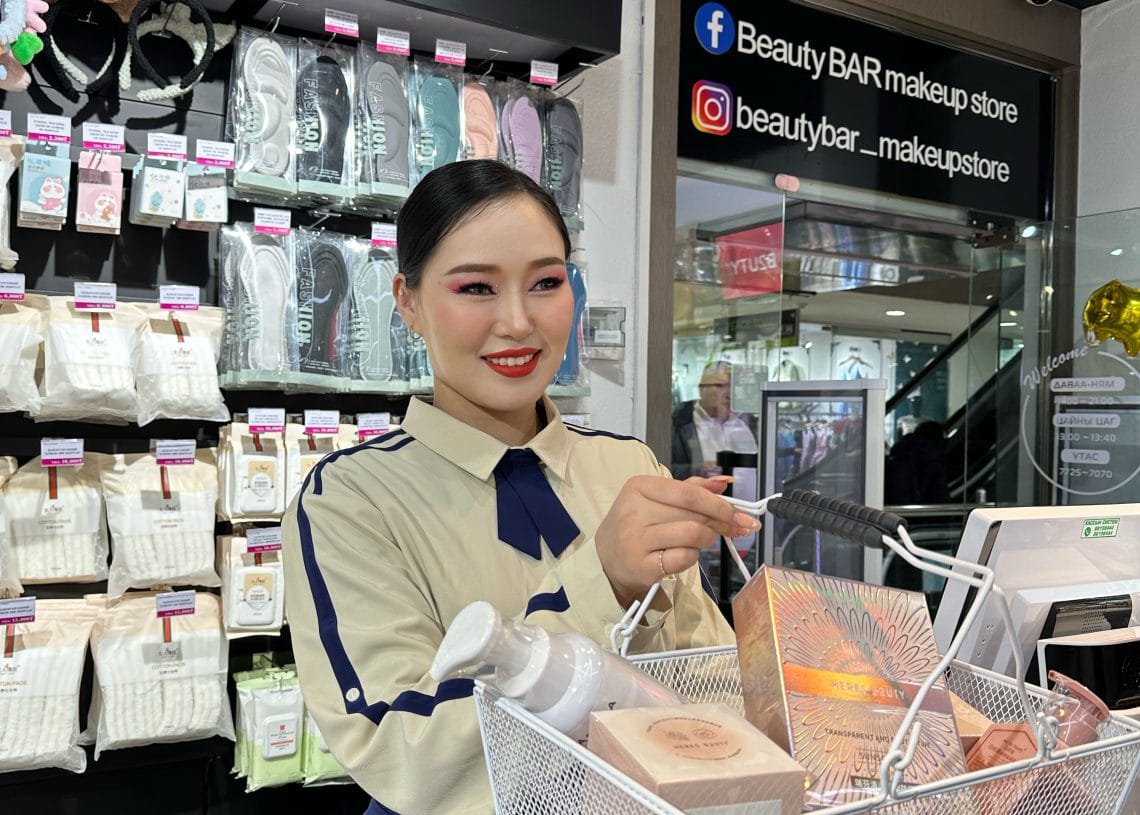 A smiling woman in a beige uniform stands behind a makeup store counter, handing a shopping basket filled with beauty products to a customer. Shelves with beauty tools and products are seen in the background.