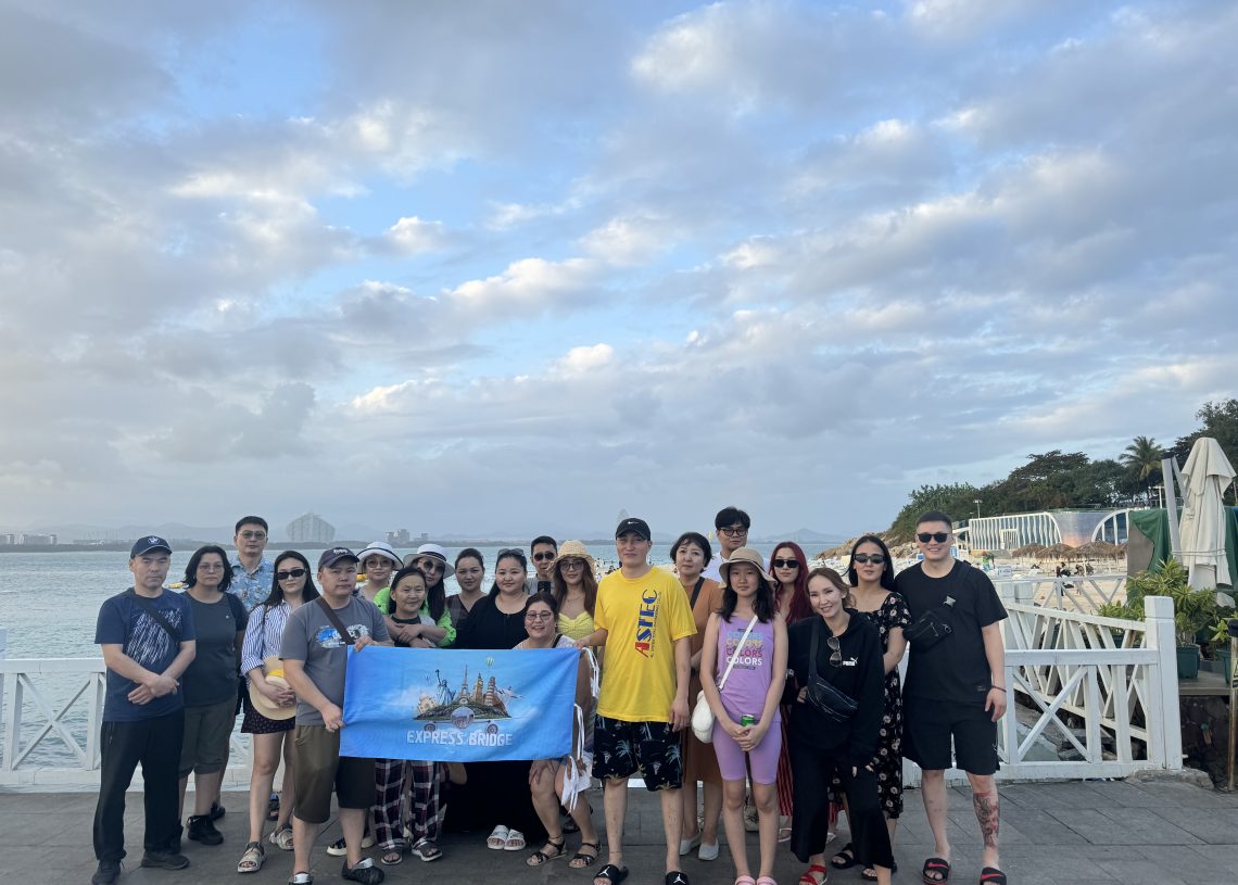 A group of people posing together outdoors by the waterfront under a partly cloudy sky. They are standing on a tiled patio, some holding a blue banner that reads "EXPRESS CHOICE." Beach chairs and umbrellas are visible in the background.