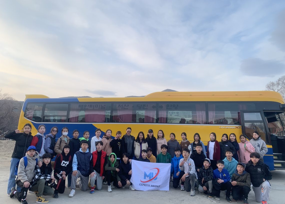 A large group of people, including children and adults, pose together in front of a yellow and blue bus. Some are holding a white flag with a colorful logo and "JPTNS BRIDGE" written on it. The sky is partly cloudy.