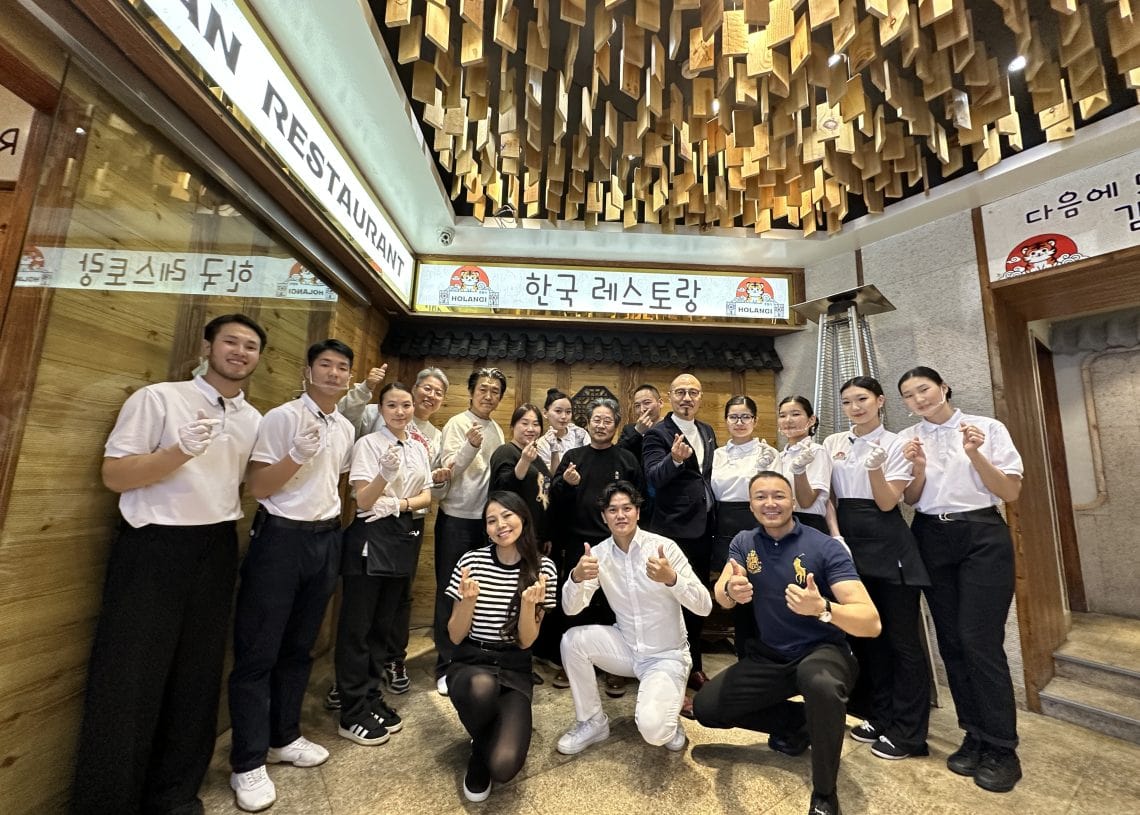 A group of people, including restaurant staff in white uniforms and others in casual clothing, pose smiling and giving thumbs up inside a Korean restaurant with wooden decor and Korean signs.