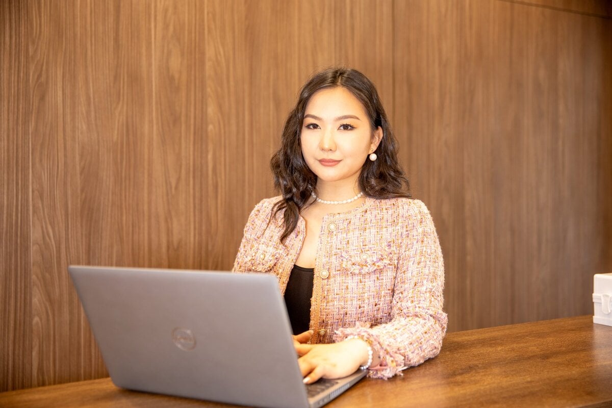 A woman with long dark hair sits at a wooden desk using a laptop. She wears a pink tweed jacket, a pearl necklace, and earrings. The background features wood-paneled walls.