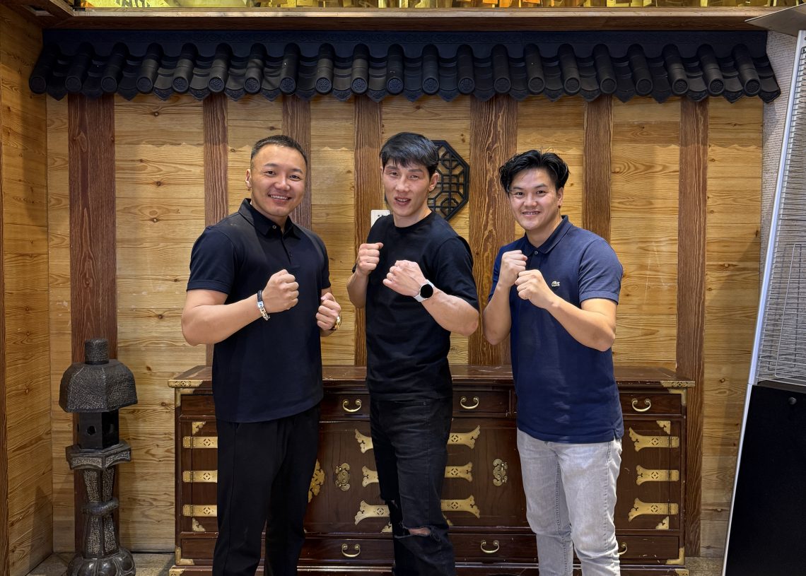 Three men stand in fighting poses, smiling, in front of a wooden wall and chest with a sign reading "Korean Restaurant" in Korean and English, and cartoon tiger logos labeled "HOLANGI.