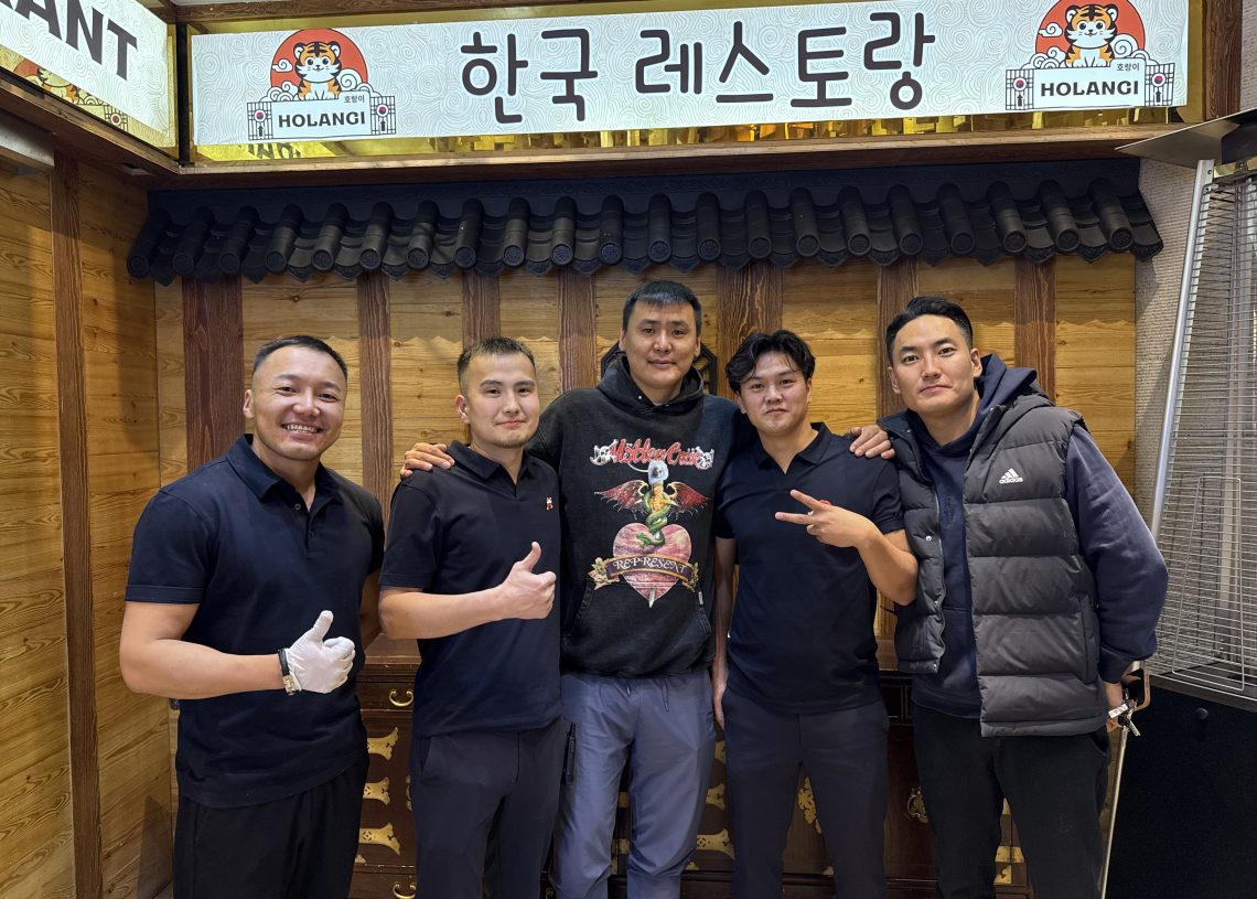 Five men stand together smiling in front of a wooden wall with a sign in Korean and English above them. Four wear black shirts, one wears a dark hoodie. They appear to be in a restaurant setting.