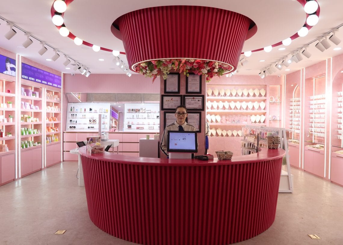 A person stands behind a bright pink reception desk in a modern, well-lit cosmetics store with shelves of colorful products and floral decorations above the counter.