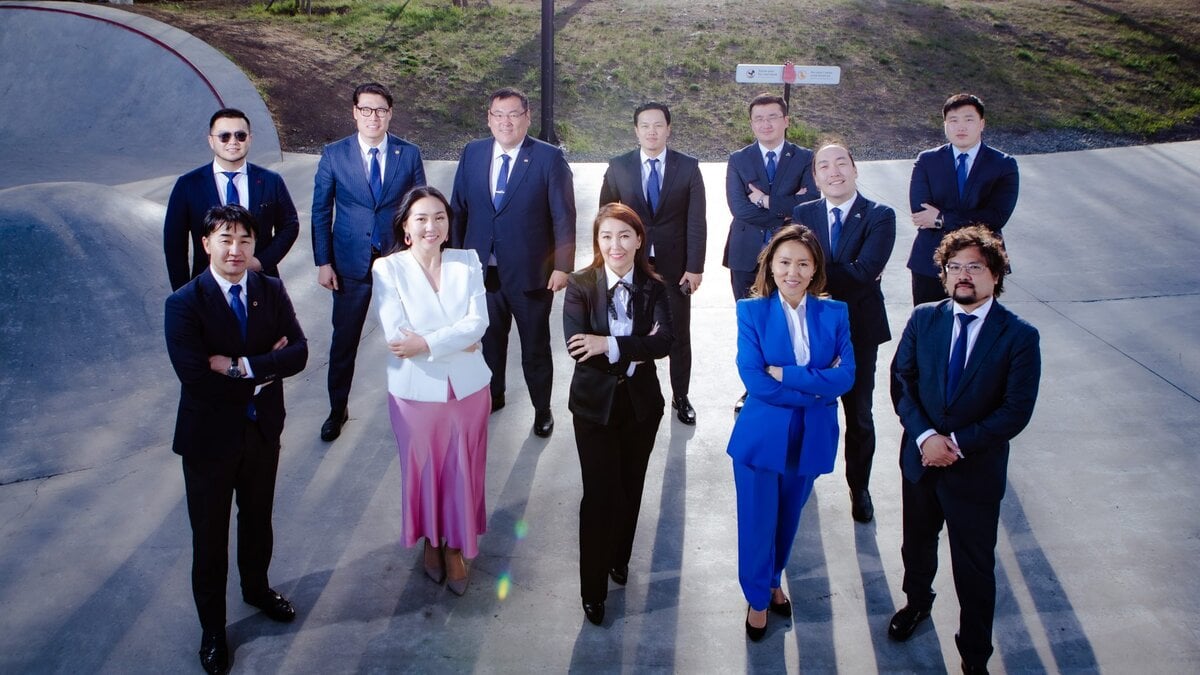A group of eleven people dressed in formal business attire stand outside on a concrete surface, smiling and posing for a photo, with sunlight casting shadows and greenery visible in the background.