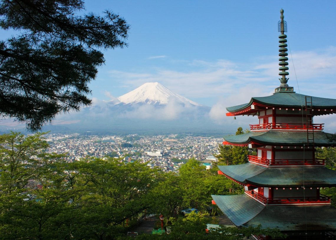 A red and green five-story pagoda stands among trees with a city below. Mount Fuji, capped with snow, rises majestically in the background under a blue sky with scattered clouds.