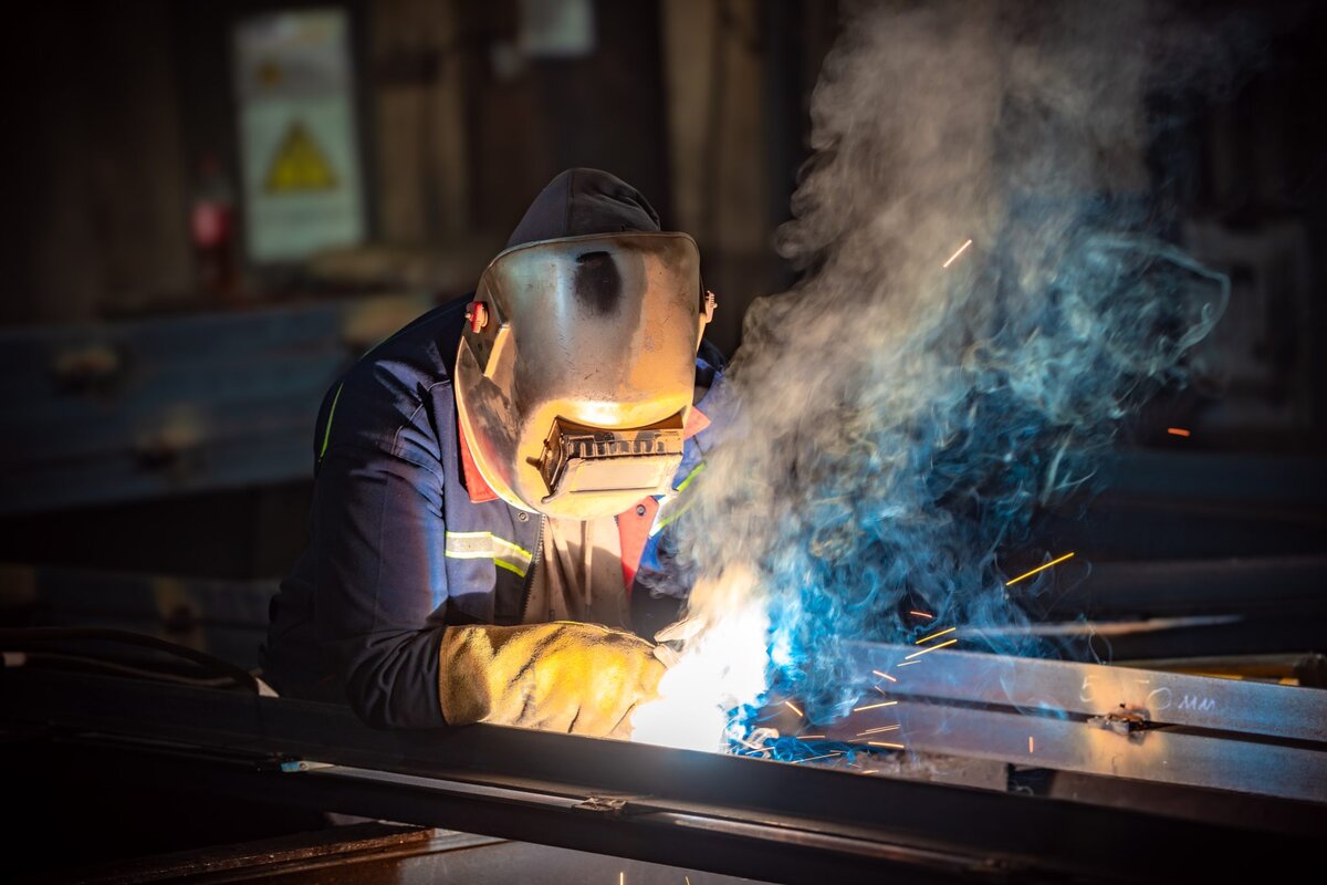 A person wearing protective gear and a welding helmet is welding metal, with bright sparks and smoke surrounding the work area in a dimly lit industrial setting.