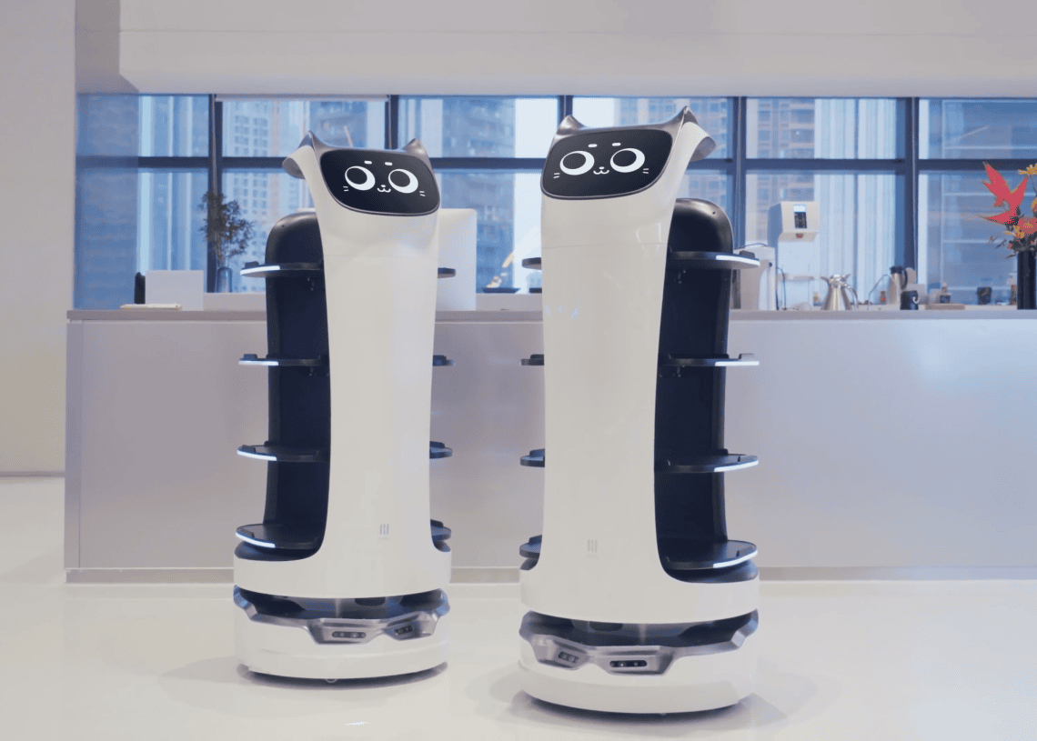 Two white, futuristic service robots with digital faces and multiple shelves stand side by side in a modern, bright room with large windows and a counter in the background.