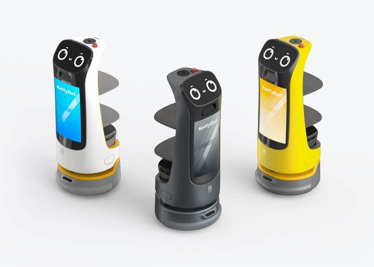 Three robot assistants with cartoon-like faces and screens stand on circular bases. Each robot is a different color: white, black, and yellow. They have multiple shelves and display the name "KettyBot" on their screens.