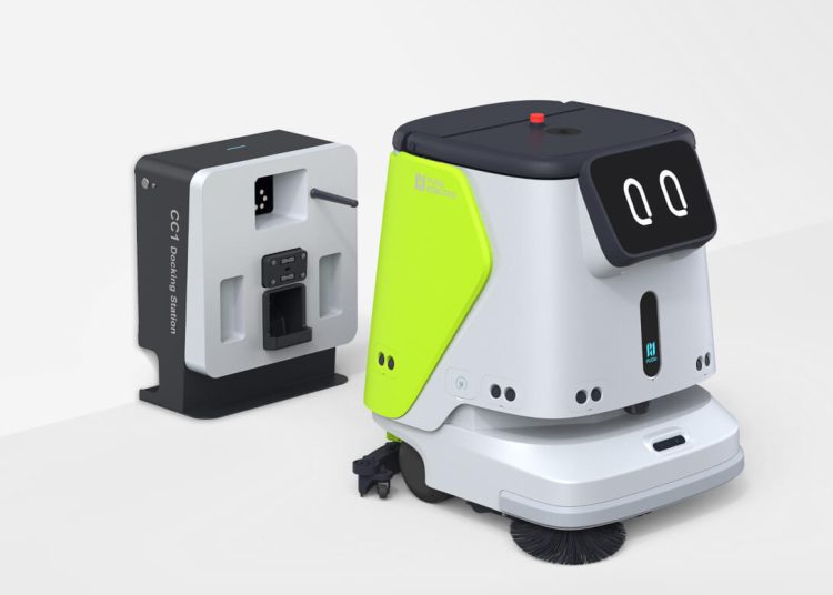 A compact floor cleaning robot with a friendly digital face is positioned next to its docking and charging station. The robot features a white and lime green body with visible brushes underneath.