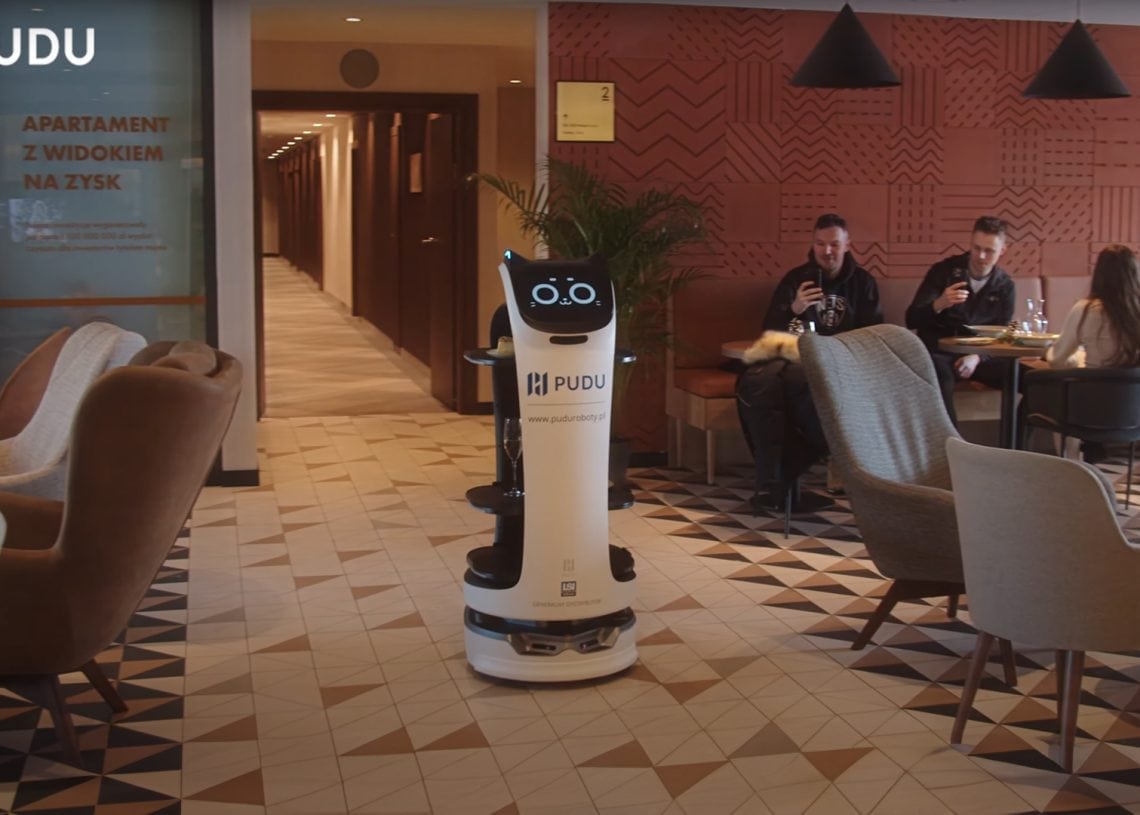 A white service robot with a display screen stands in a modern cafe lounge, while people sit at tables chatting and using their phones. The decor features patterned flooring and warm-toned walls.