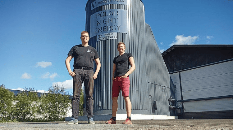 Two people stand in front of a large, gray industrial structure labeled "Polar Night Energy." The sky is clear and blue, and there is a building and some greenery nearby.