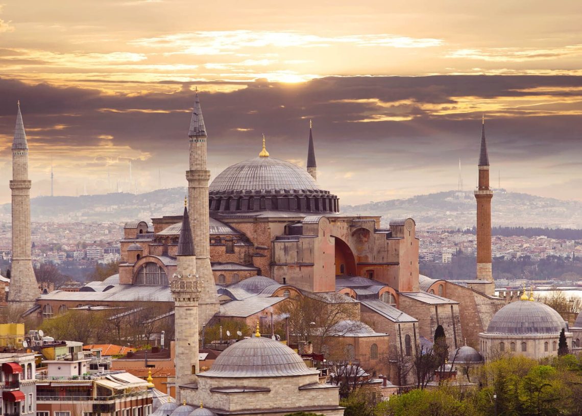 The Hagia Sophia in Istanbul, Turkey, with its domes and minarets, is seen at sunset under a dramatic sky, surrounded by city buildings and greenery.