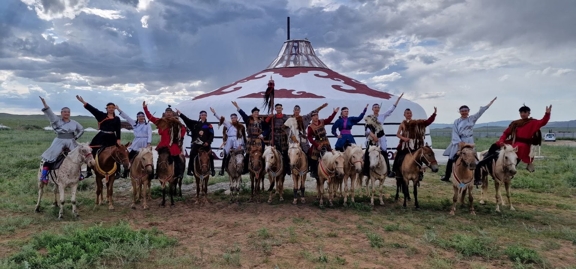 A group of people in traditional clothing pose on horseback, raising one arm, in front of a large, decorated yurt in a grassy field under a cloudy sky.