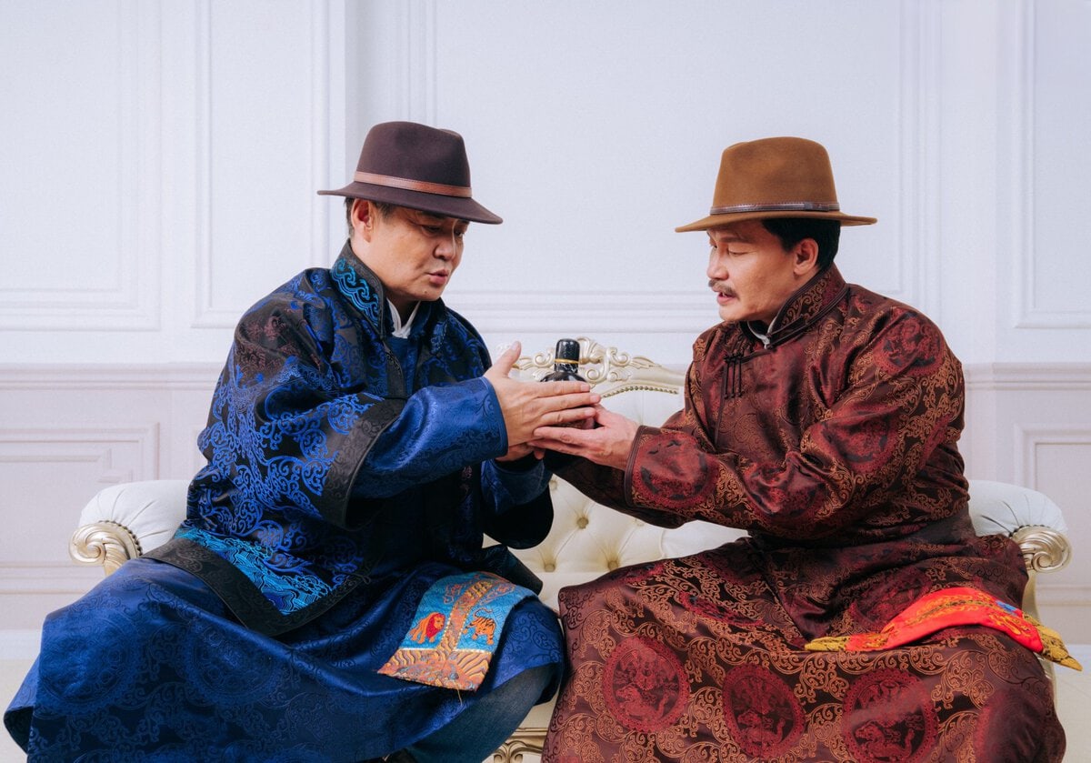 Two men in ornate traditional robes and hats sit on a white sofa, holding a small ornate cup or bowl between them, appearing to share a ceremonial or meaningful moment.