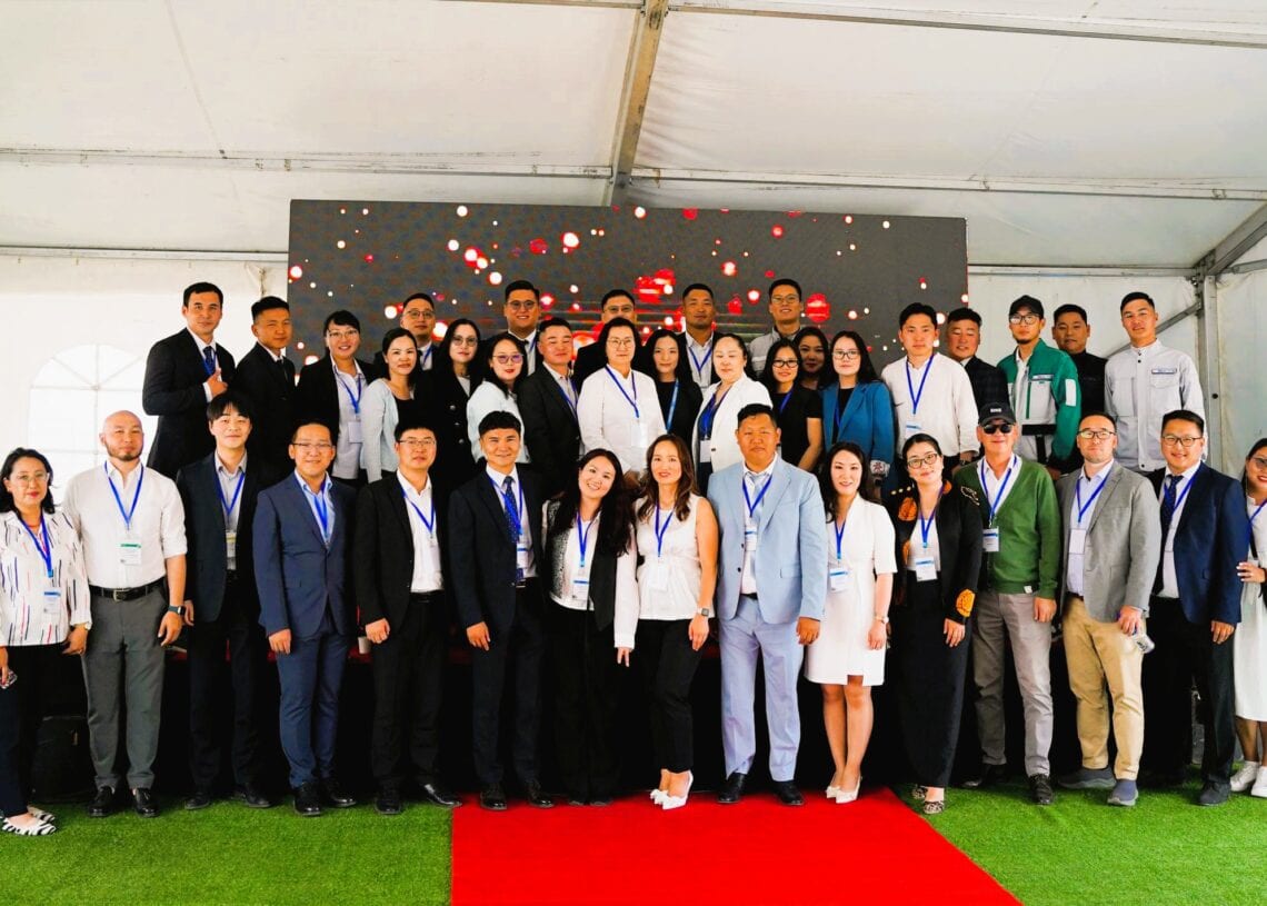 Нэрийн тэмдэгтэй ажил хэрэгч хувцастай хүмүүс ногоон хиймэл зүлгэн дээрх цагаан майхан дор цугларч, ардаа дижитал дэлгэцтэй улаан хивсний дээгүүр зогсож, ногоон барилга, ногоон ирээдүйгээ тэмдэглэж байна. - Business.MN