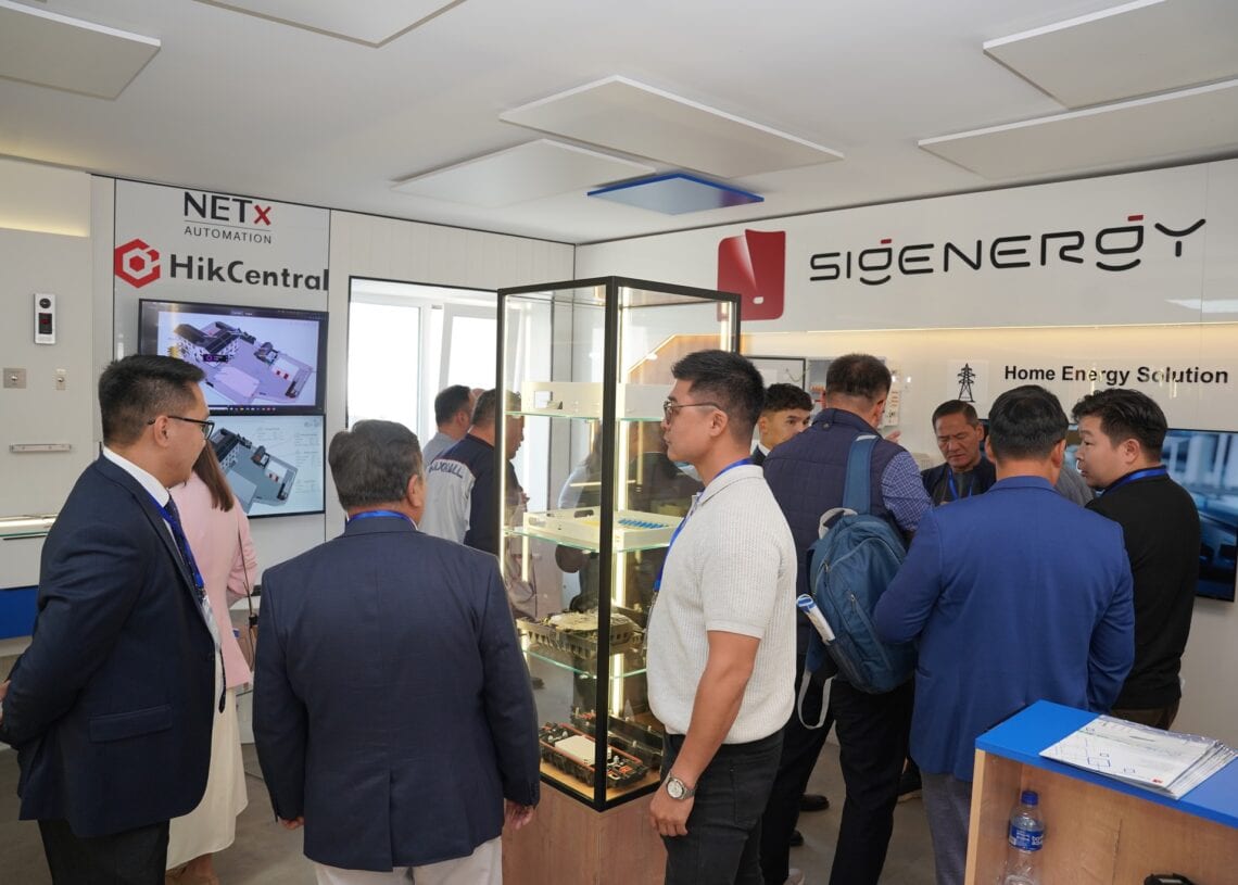 Хэсэг хүмүүс орчин үеийн үзэсгэлэнгийн танхимд электрон эд анги бүхий дэлгэцийн хайрцагны эргэн тойронд зогсож байна. SIGENERGY болон HikCentral брэндүүд ханан дээр харагдаж байгаа нь ухаалаг барилга, ногоон ирээдүйн шинэлэг санааг онцолжээ. Зарим оролцогчид хурлын тэмдэг зүүдэг. - Business.MN