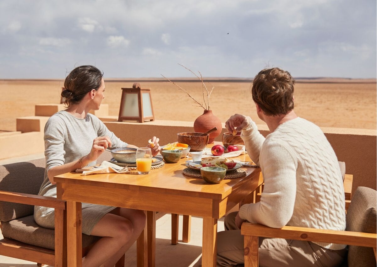 Хоёр хүн модон ширээний ард суугаад гадаа өглөөний цайгаа ууж, цөлийн ландшафтыг харан зогсох нь тогтвортой аялал жуулчлалын төгс орчин юм. Ширээн дээр аяга, тавагтай хоол, шүүс, шавар савны гол хэсэг байдаг бөгөөд тэд үзэмжийг биширдэг. - Business.MN