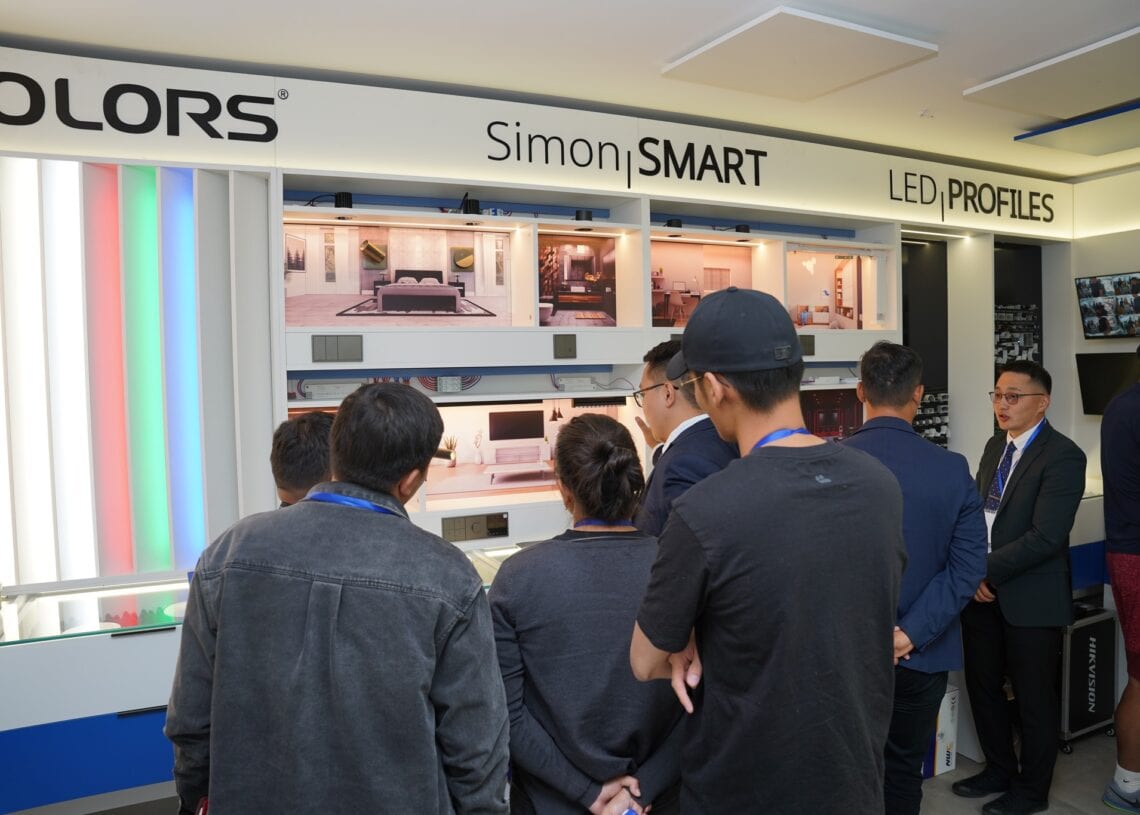 Хэсэг хүмүүс LED гэрэлтүүлгийн бүтээгдэхүүн, ухаалаг гэрийн тохиргоог харуулсан үзүүлэнгийн өмнө зогсож, эдгээр шинэлэг зүйл ногоон барилга, ухаалаг барилга шийдэлд хэрхэн хувь нэмэр оруулж байгааг илтгэгч тайлбарлаж байна. - Business.MN