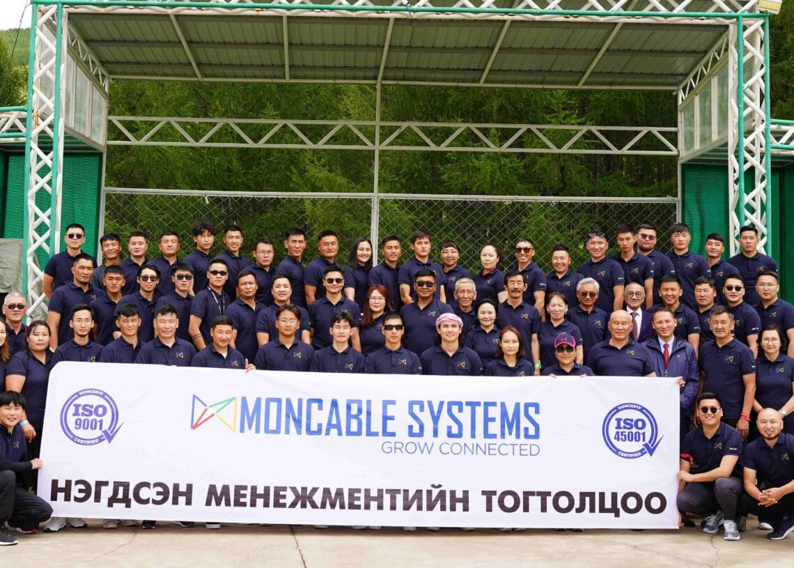 Тэнгисийн цэргийн цамц өмссөн томоохон бүлэг ISO лого, монгол бичвэр бүхий “Монкабле системс”-ийн туг барин гадаа зургаа авахуулж, ногоон дов толгод, моддын дунд ухаалаг барилгыг тэмдэглэж, ногоон ирээдүйн төлөө зүтгэж буйгаа илэрхийлж байна. - Business.MN