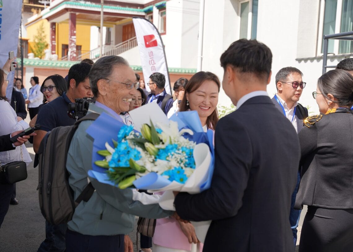 Инээж буй хүмүүс, гэрэл зурагчдын дунд, цаана нь ногоон ирээдүйн сурталчилгаа, орчин үеийн ухаалаг барилга бүхий гадаа өөр эрэгтэйгээс том баглаа цэнхэр, цагаан цэцгийн баглааг эмэгтэй, эрэгтэй хоёр баяртайгаар хүлээн авч байна. - Business.MN