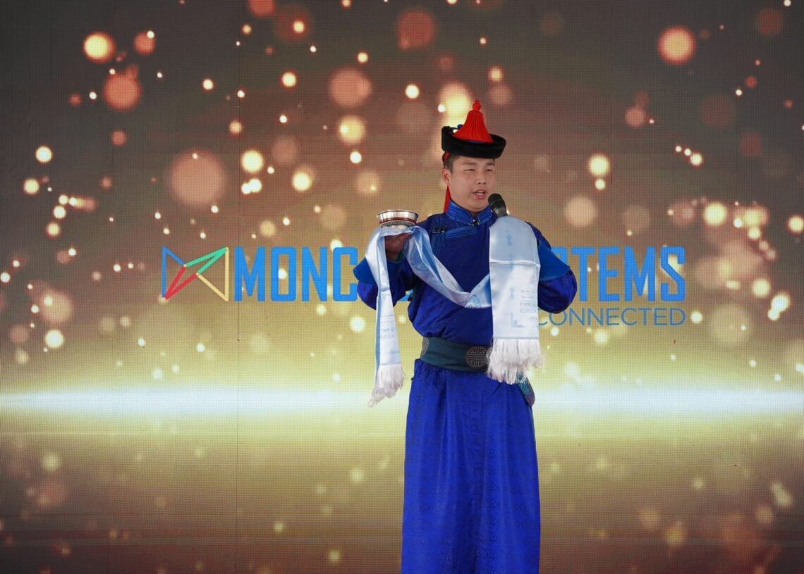 Монгол үндэсний хувцас, улаан малгай өмссөн хүн тайзан дээр гялалзсан гэрэл болон "MONG SYSTEMS" гэсэн хэсэгчилсэн бичвэрийн дор ёслолын цагаан ороолт барьж, ухаалаг барилга, ногоон ирээдүйг тэмдэглэж байна. - Business.MN