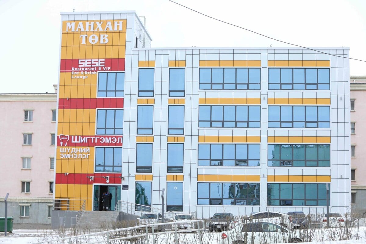Орчин үеийн дөрвөн давхар шар, цагаан, улбар шар өнгийн фасадтай, том цонхтой, монгол бичгээр бичсэн тэмдэг бүхий орчин үеийн барилга. Урд талд нь машинууд зогсож, газар цасанд дарагдана. - Business.MN