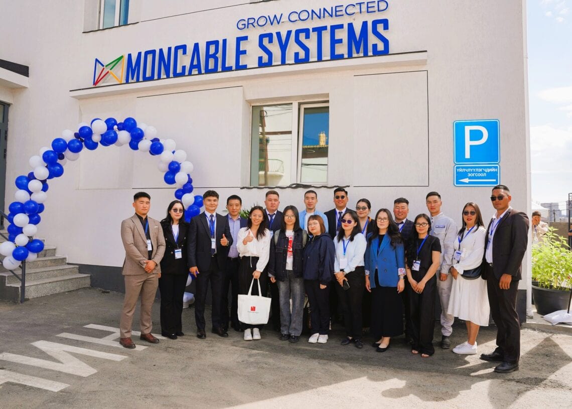 Цэнхэр цагаан бөмбөлөг нуман хаалганы хажууд "Монкабл системс" гэсэн бичиг наасан барилгын гадаа хэсэг хүмүүс зургаа авахуулж байна. Ихэнх нь ухаалаг барилга, ногоон ирээдүйгээ тэмдэглэж, ажил хэрэгч хувцас өмсөж, инээмсэглэж байна. - Business.MN